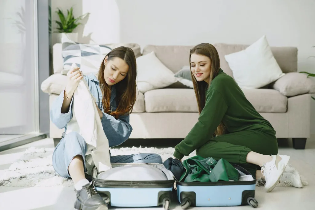 Optimistic young female travelers in trendy outfits sitting on floor against comfortable sofa and unpacking suitcase in modern hotel room