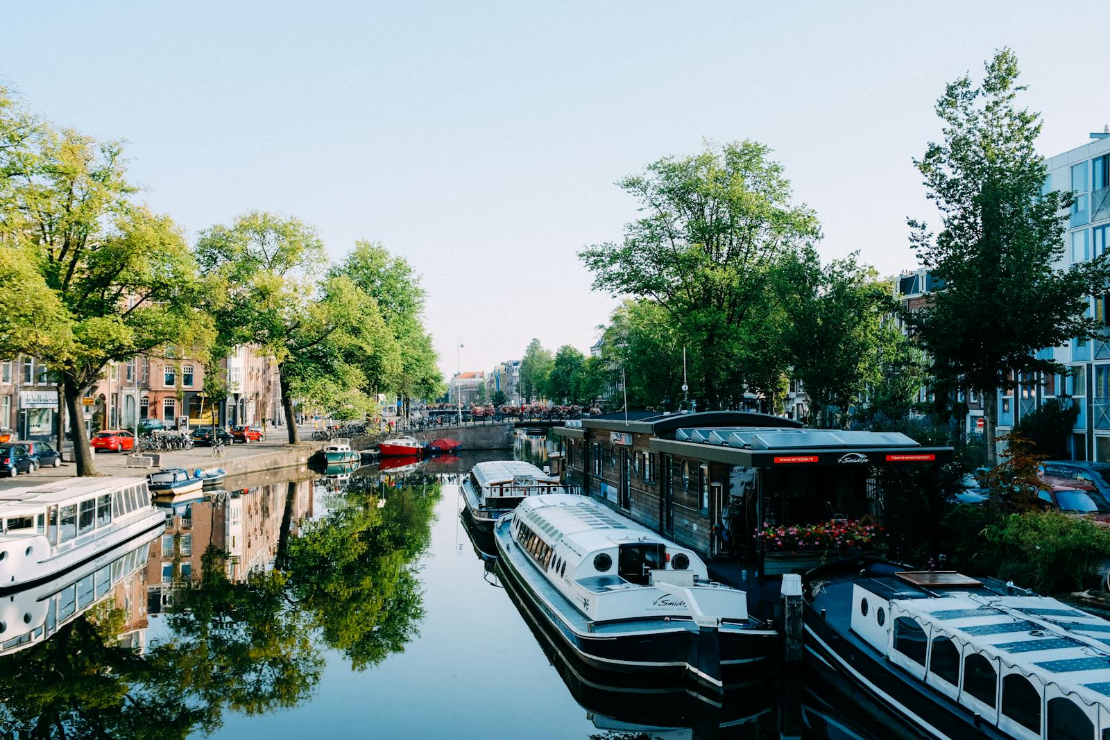 Modern cruise boats moored on calm canal reflecting green trees and buildings on sunny day